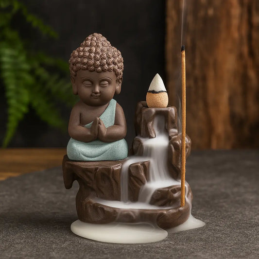 Brûleur d’encens en céramique représentant un bébé Bouddha en méditation avec cascade à reflux, décoré d’un cône et d’un bâton d’encens sur fond ardoise et bois clair.