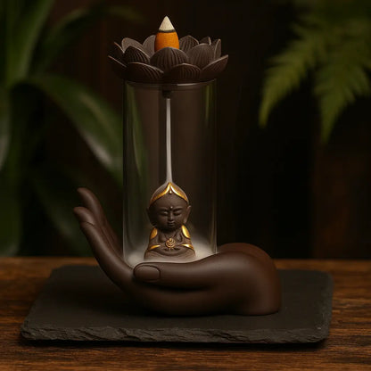Fontaine d’encens en forme de main tenant un Bouddha doré sous cloche en verre, sur ardoise sombre avec fond bois et végétal chaleureux