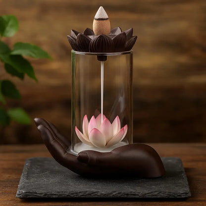 Fontaine d’encens en forme de main tenant un lotus rose sous cloche en verre, sur ardoise sombre et décor bois-végétal chaleureux